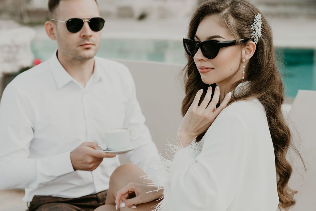 Elegant couple in white outfits and black sunglasses relaxing by a pool, woman wearing a decorative hair accessory.