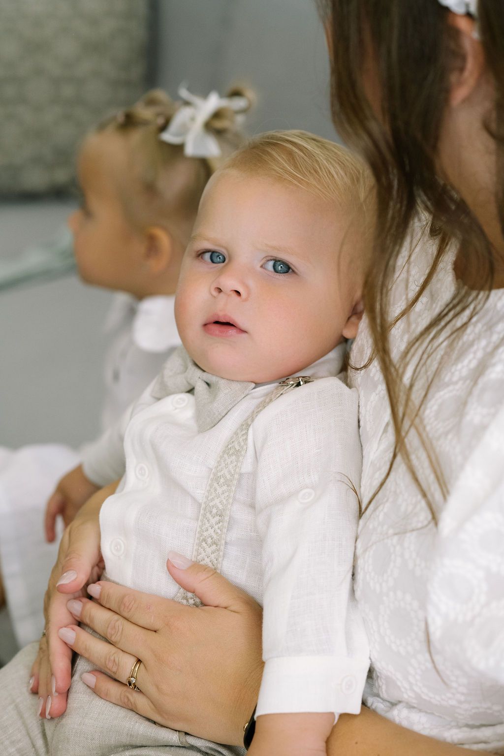 A blue-eyed baby in white linen outfit sits on mothers lap during a christening ceremony.