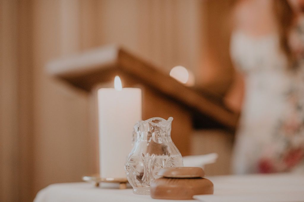 White unity candle with crystal figurine on wooden base, soft bokeh background with bride in floral dress.