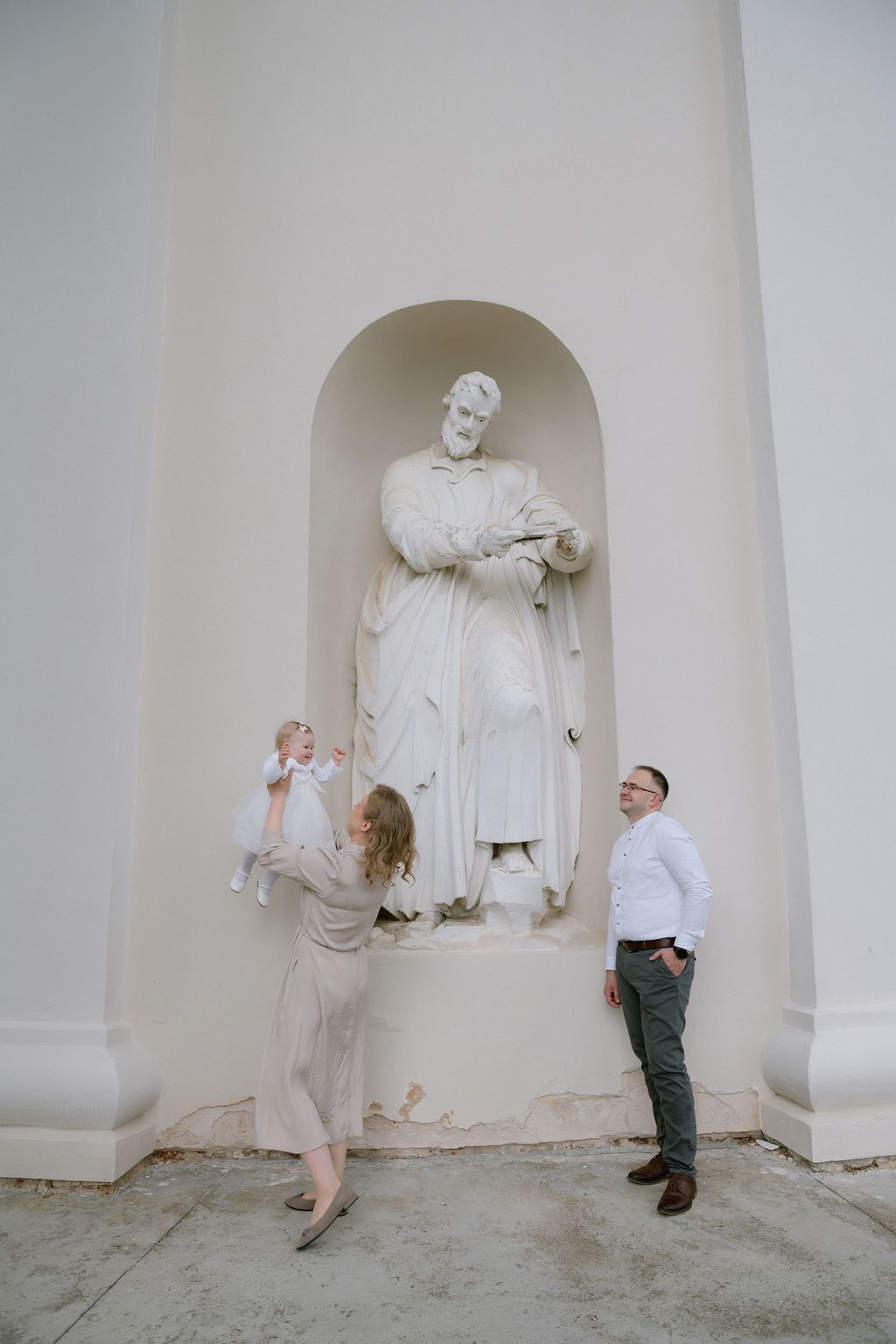 Family posing by a white classical statue in an arched niche on a white building exterior.