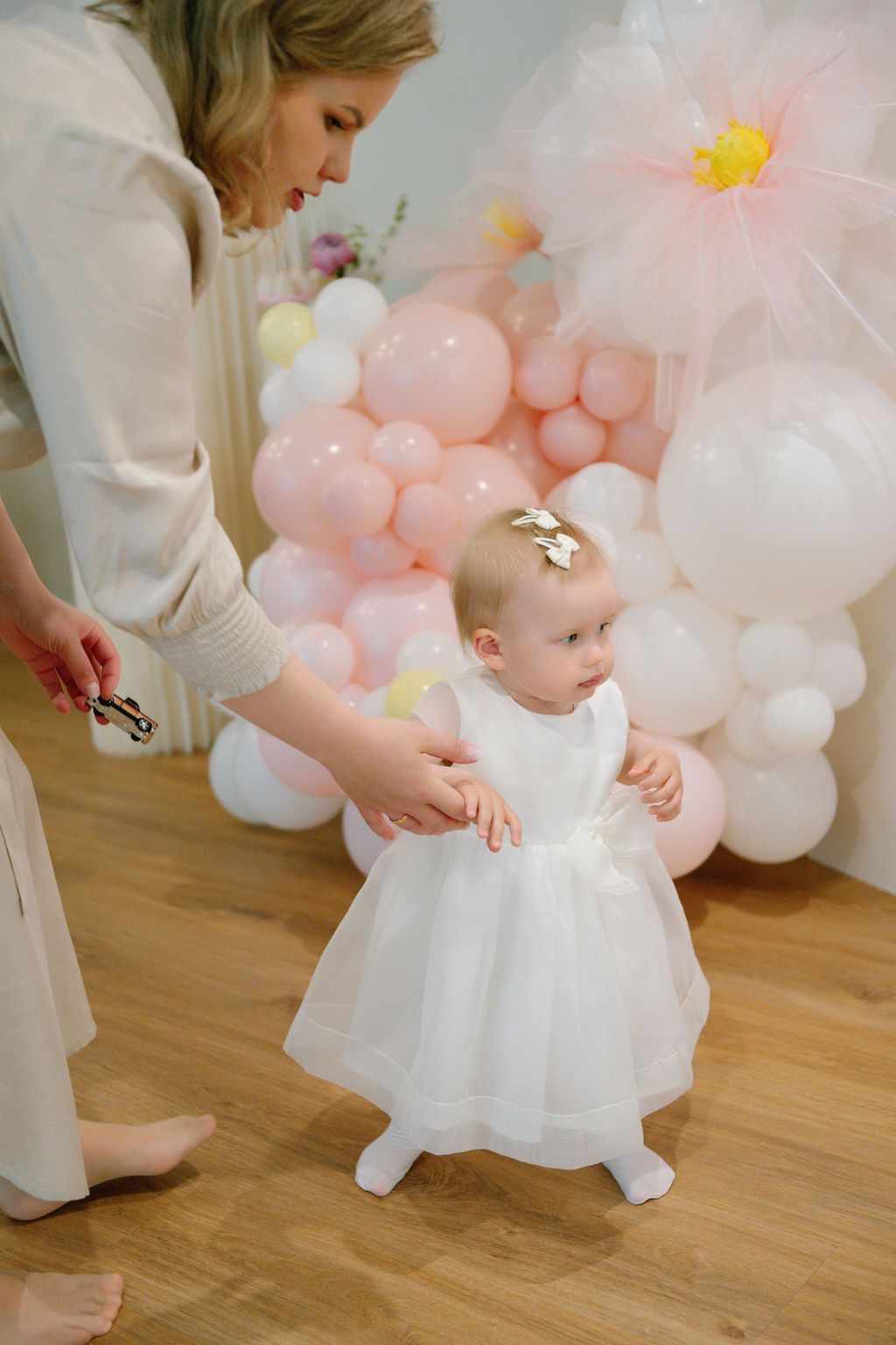 Mother helping baby girl in white dress stand at first birthday party with pink balloon decorations.