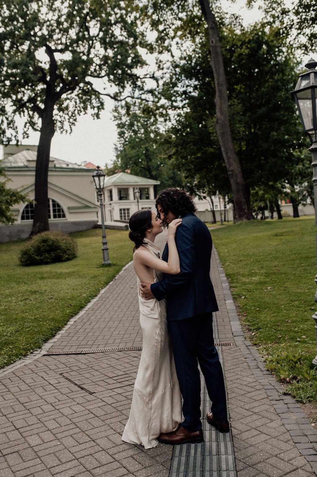 Bride and groom share a romantic kiss on a brick pathway surrounded by lush green trees and a historic building.