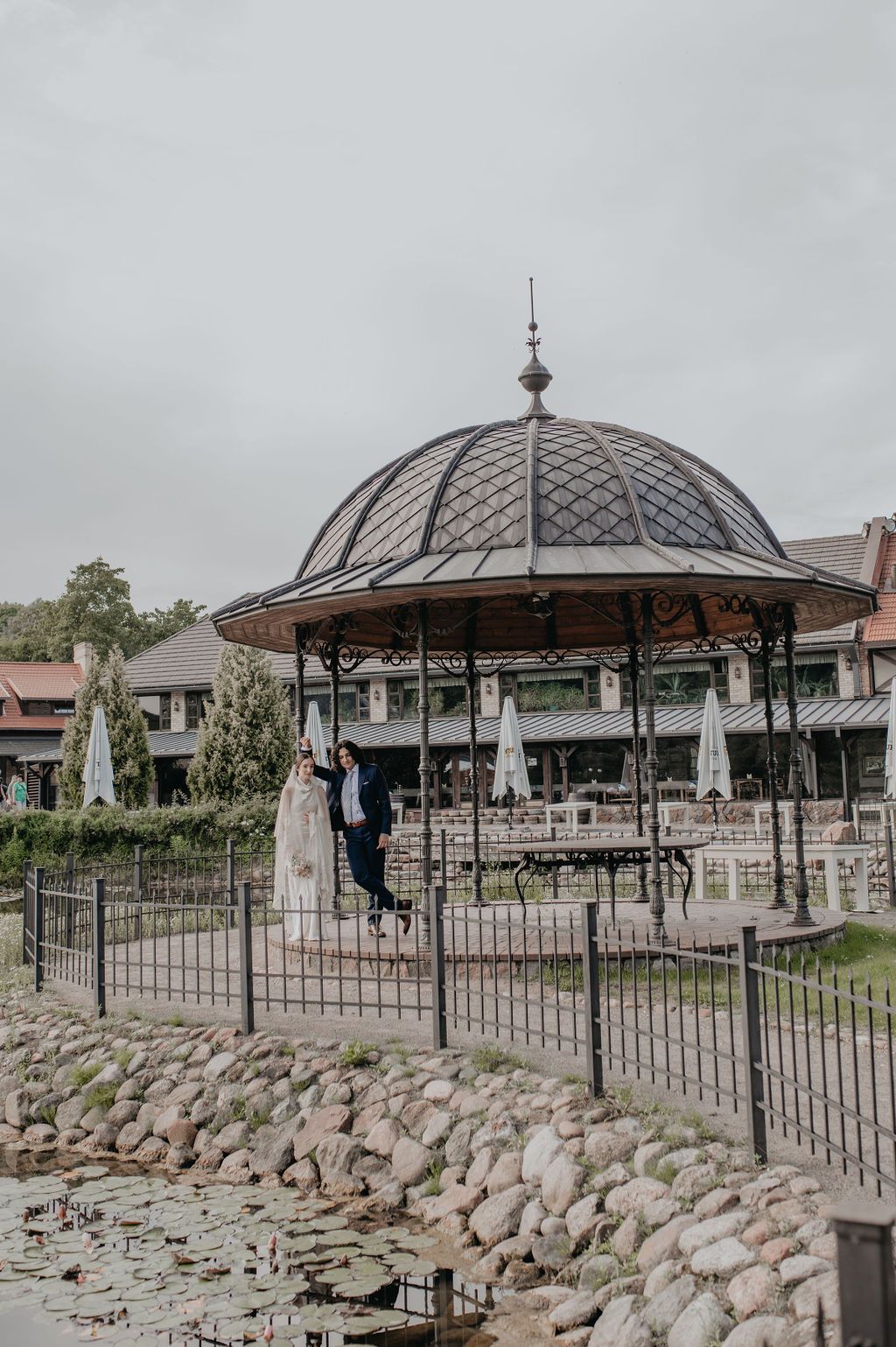 Bride and groom posing near an ornate gazebo by a lily pond with stone borders at an elegant wedding venue.