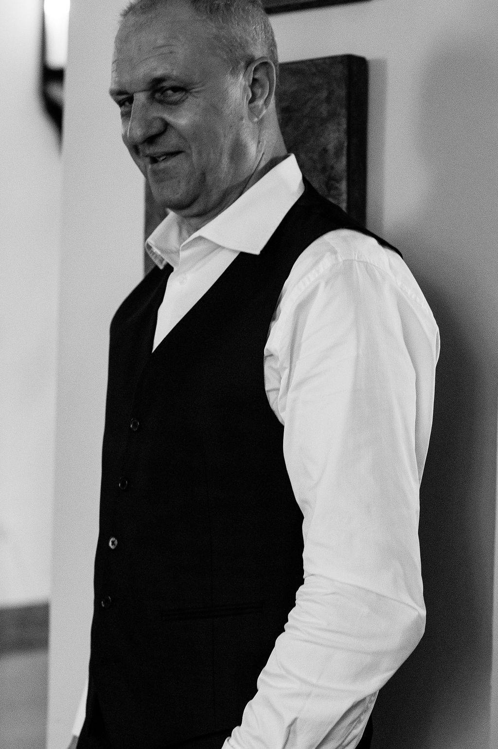 Black and white portrait of a smiling middle-aged man in a white shirt and dark vest leaning against a wall.