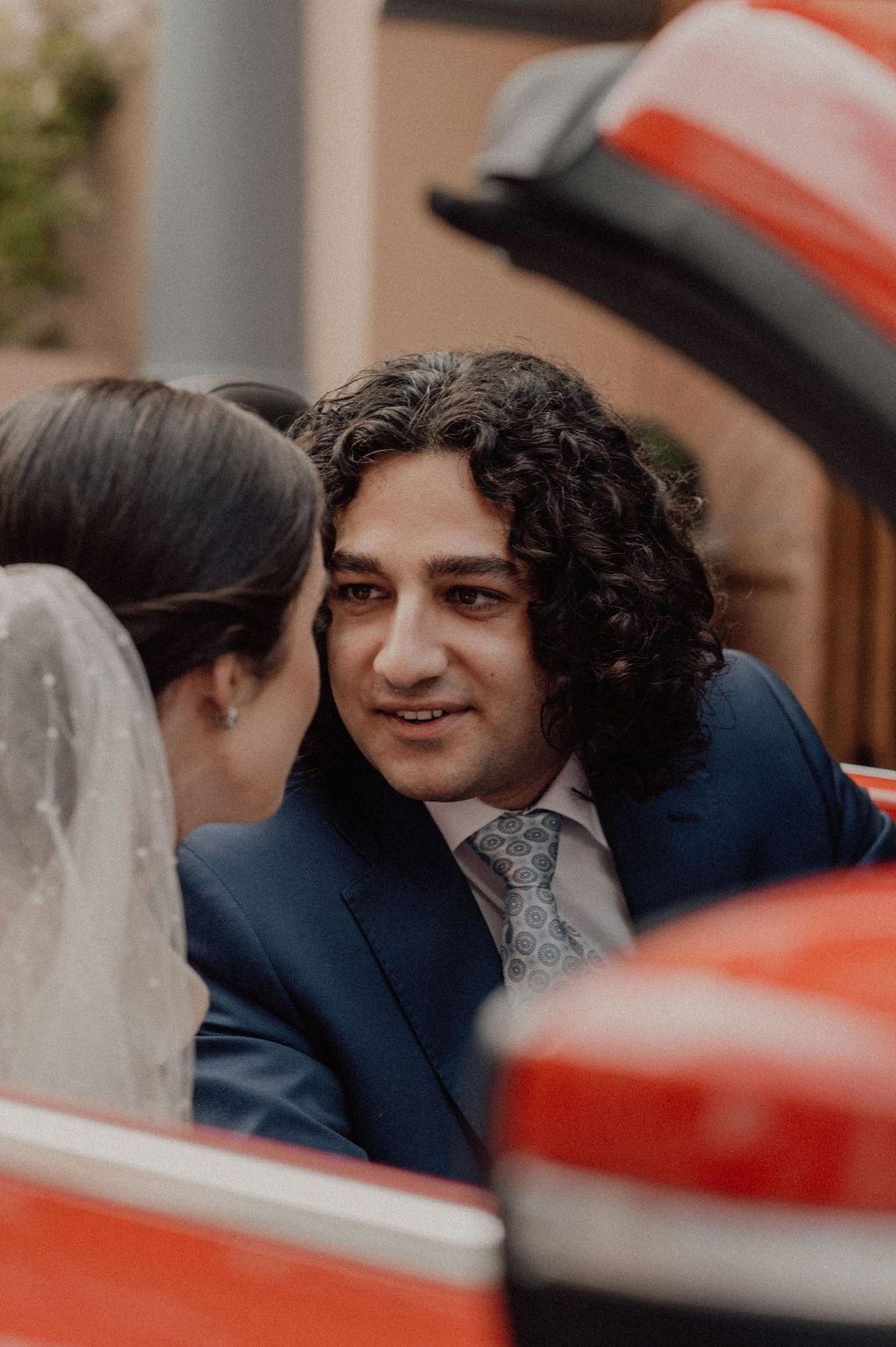 Groom with curly hair in navy suit and tie smiling at bride in white veil inside a red convertible car.