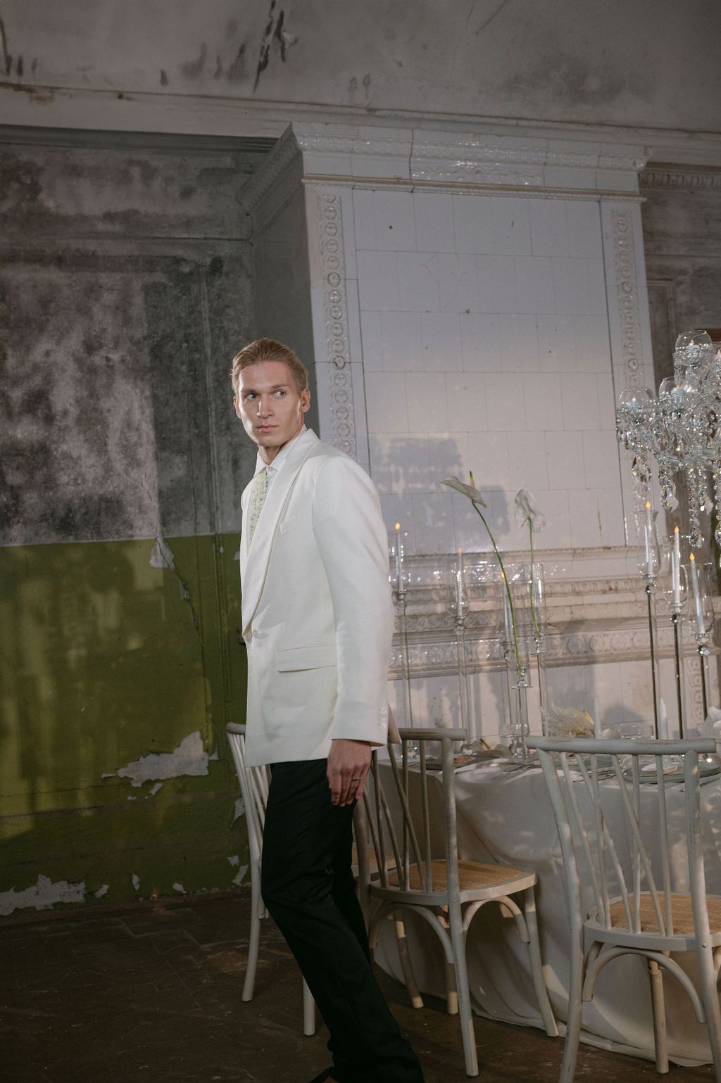 Blonde man in white blazer and black trousers stands beside crystal candelabra in rustic vintage room.