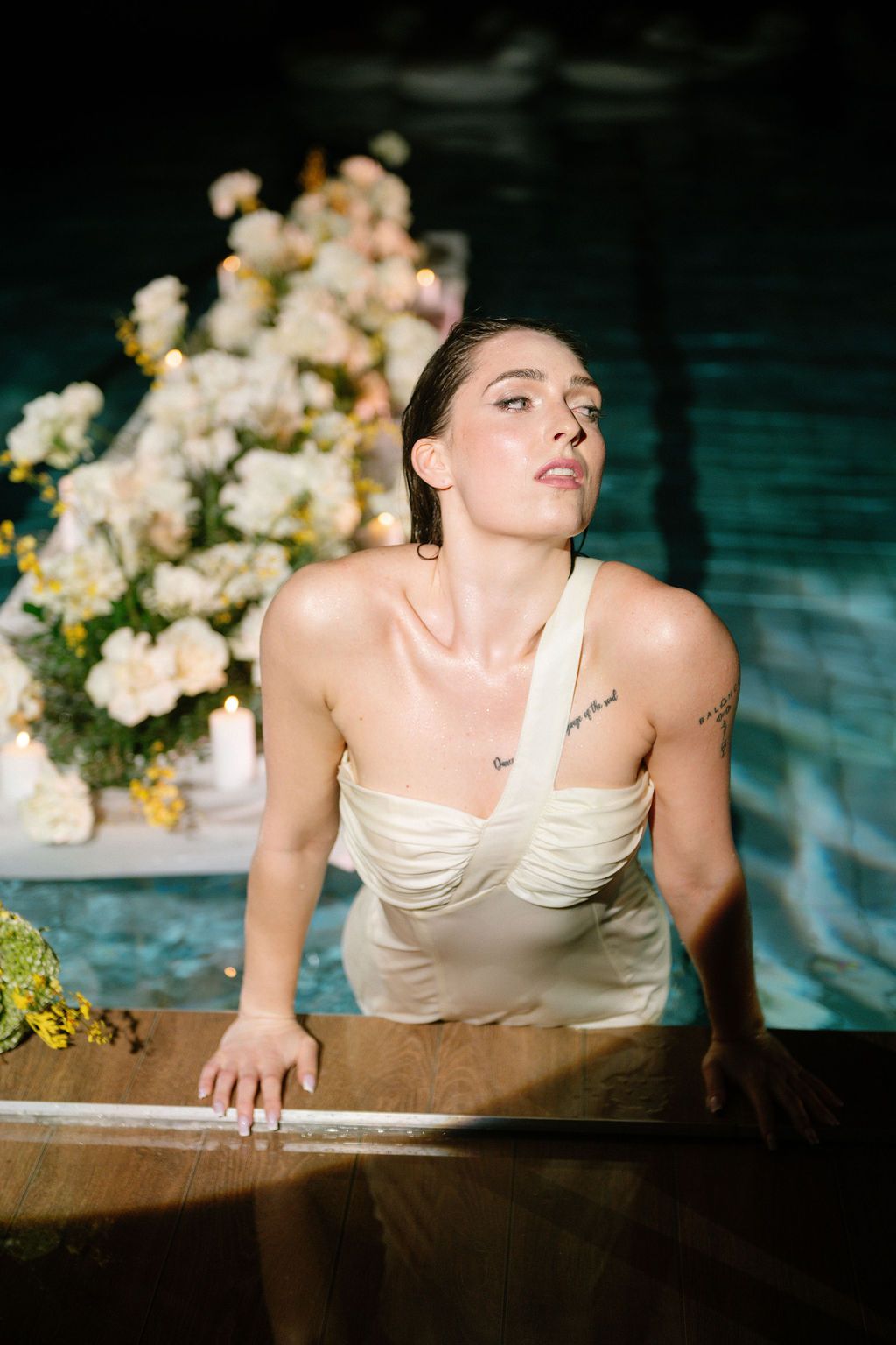 Woman in white dress emerging from pool with floral arrangements in background.