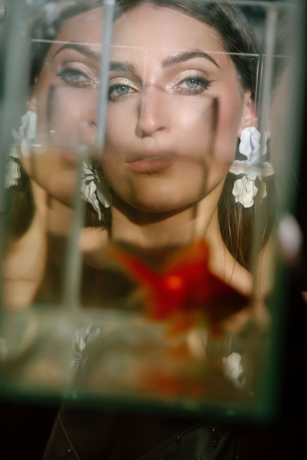 Woman with floral earrings poses behind cracked glass with a red origami star, dramatic eye makeup.