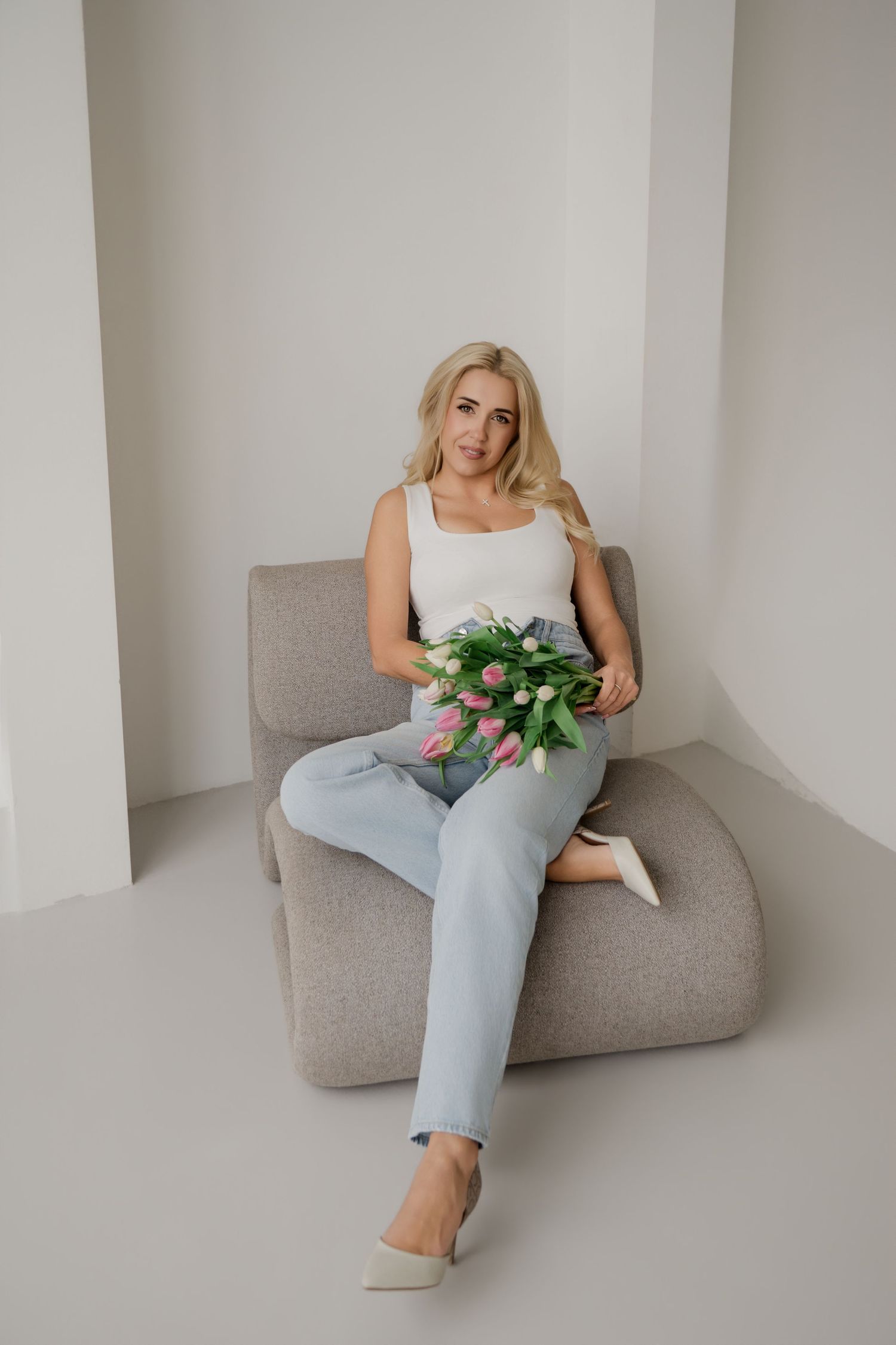 Blonde woman sitting on gray chair holding pink and white tulips, wearing white tank top and blue jeans in minimalist studio.