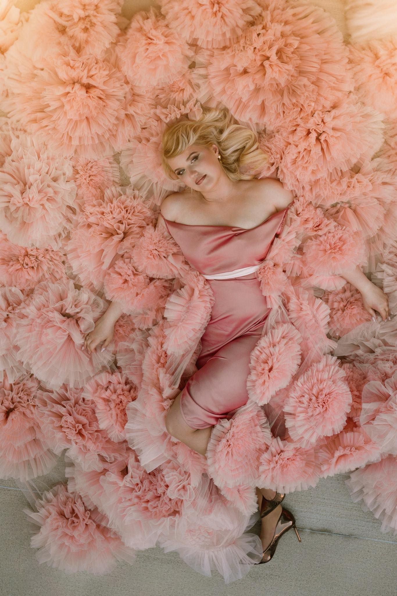 Blonde woman in pink satin dress lying among voluminous pink tulle ruffles, viewed from above in elegant fashion portrait.