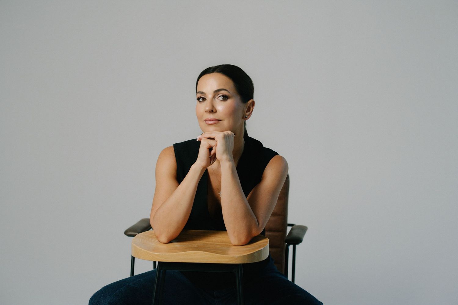 Thoughtful woman in black sleeveless top sits on wooden chair, chin resting on hands, gazing upward against gray background.