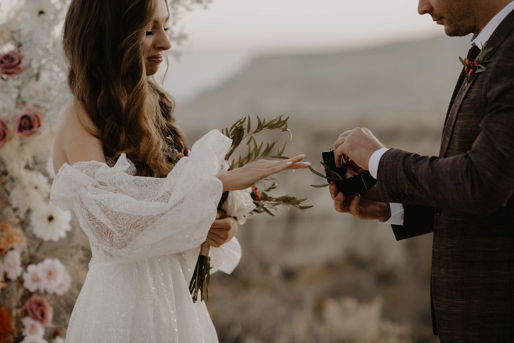 Bride in lace gown receiving ring from groom at outdoor wedding ceremony with floral backdrop.