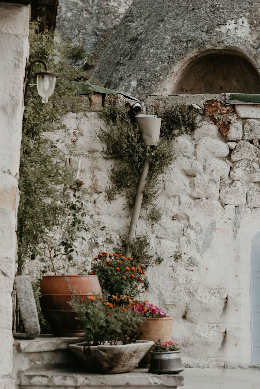 Rustic stone wall with terracotta pots of colorful flowers and a vintage lamp in a charming Mediterranean courtyard.