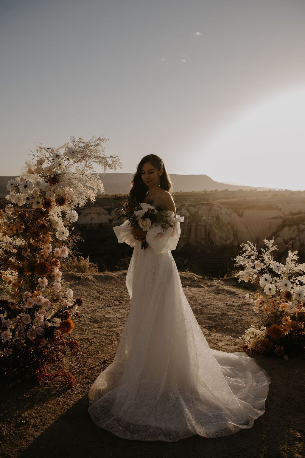 Bride in white off-shoulder gown with floral arch at golden hour desert wedding.