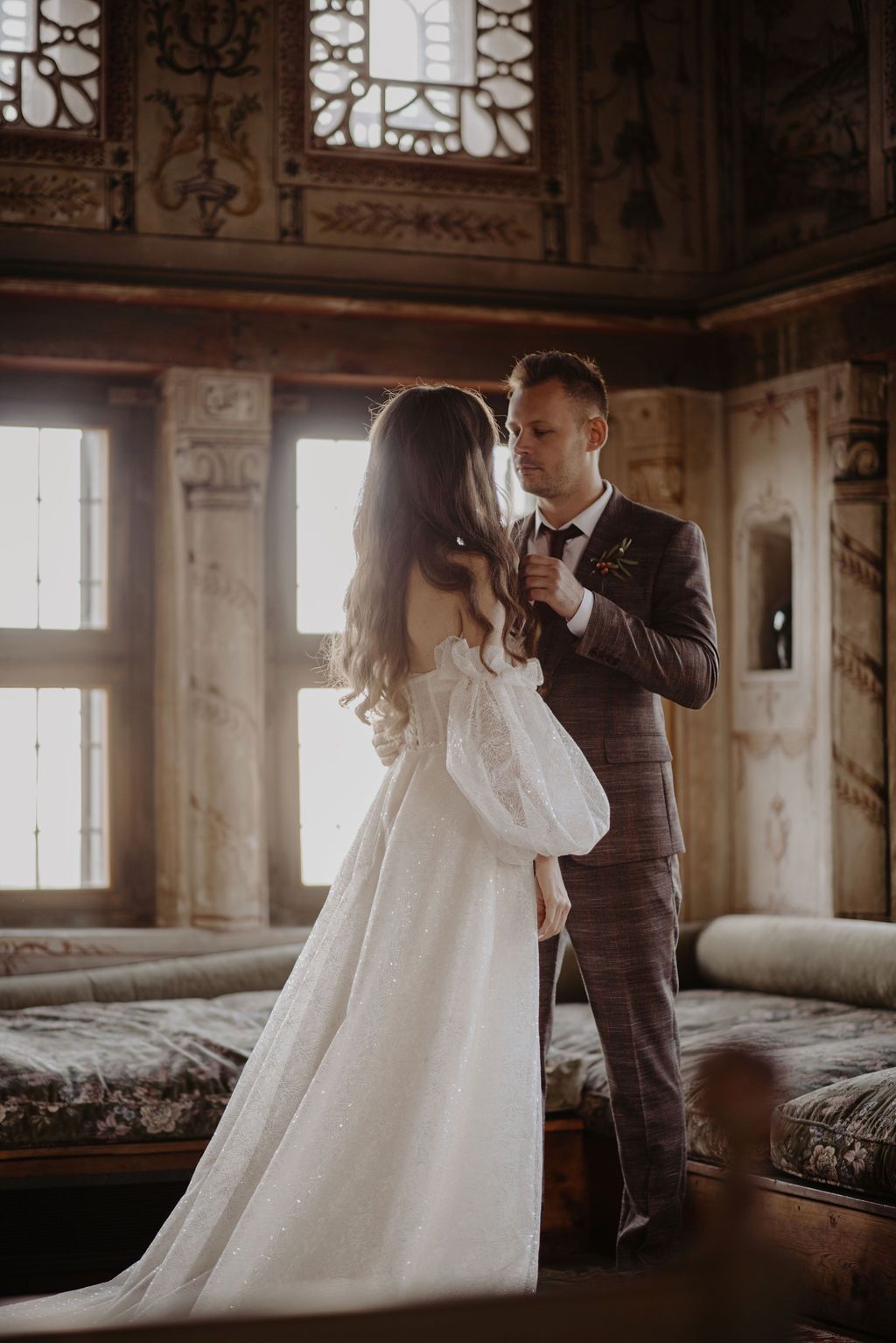 Bride and groom share a romantic moment in an ornate vintage room, bride in white gown, groom in plaid suit.