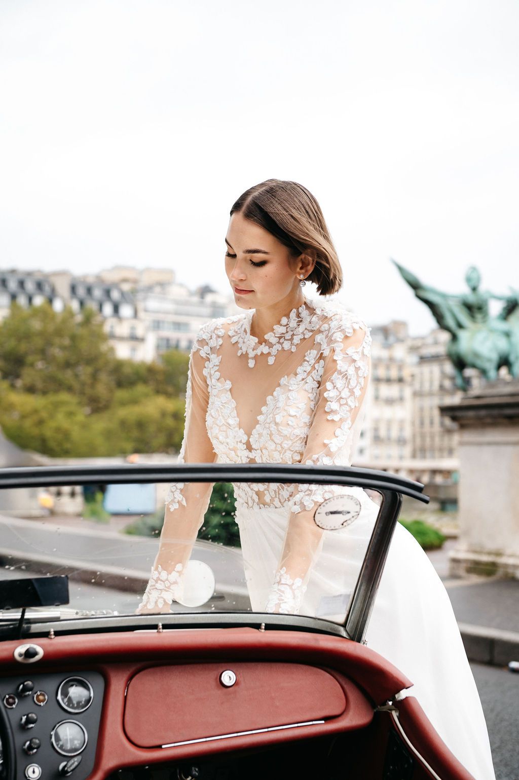 Bride in floral lace gown stepping into a vintage convertible car with Parisian architecture in the background.