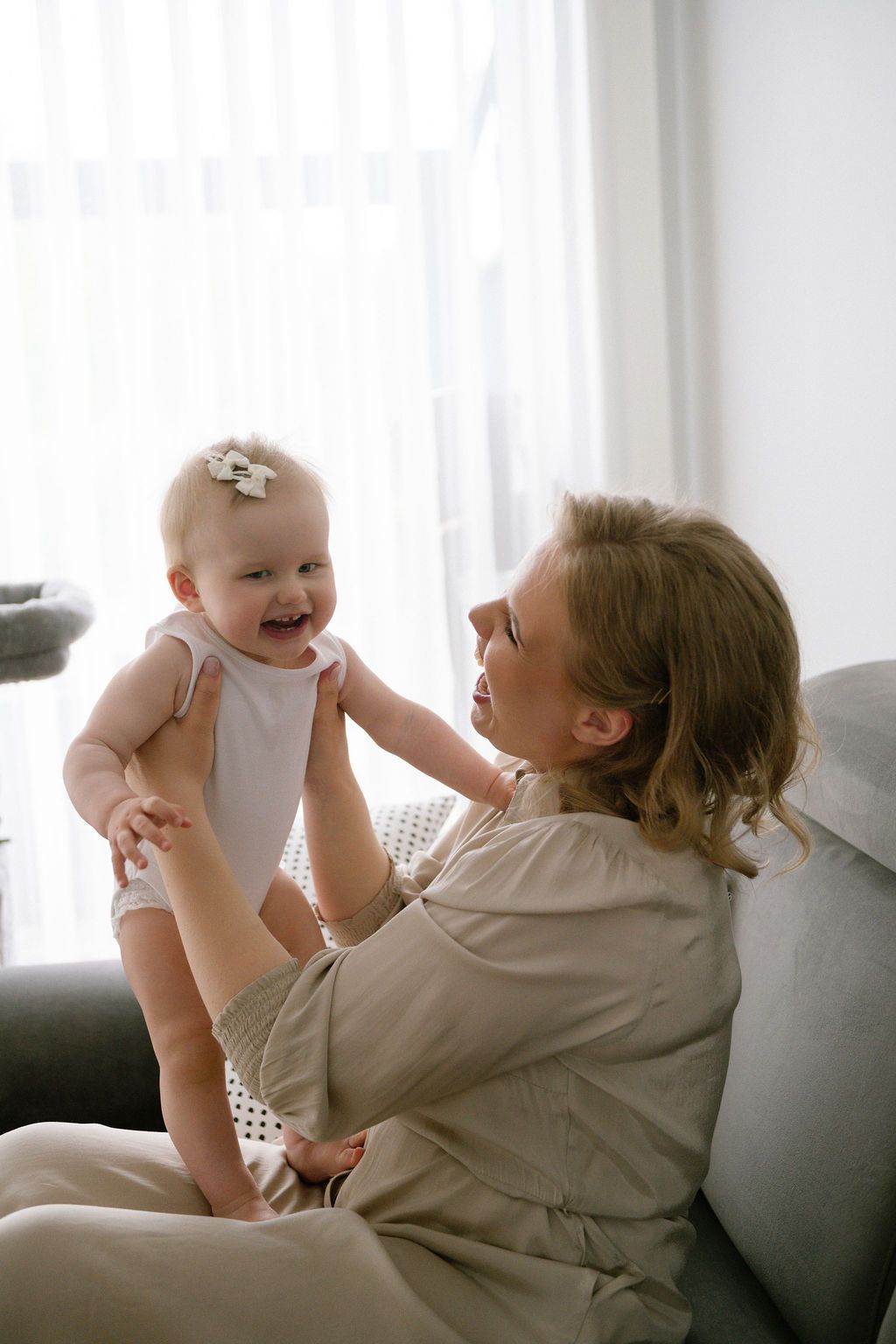A smiling mother lifts her happy baby girl in a bright living room, sharing a joyful bonding moment.