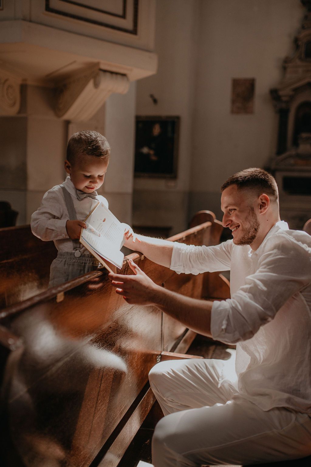 Father and toddler son reading book together on church pew, sharing a joyful moment in warm light.