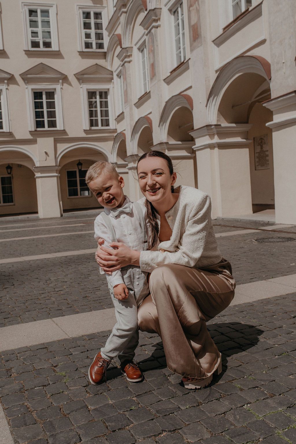 A smiling woman crouches beside a young boy in a sunny European courtyard, both dressed in elegant beige outfits.