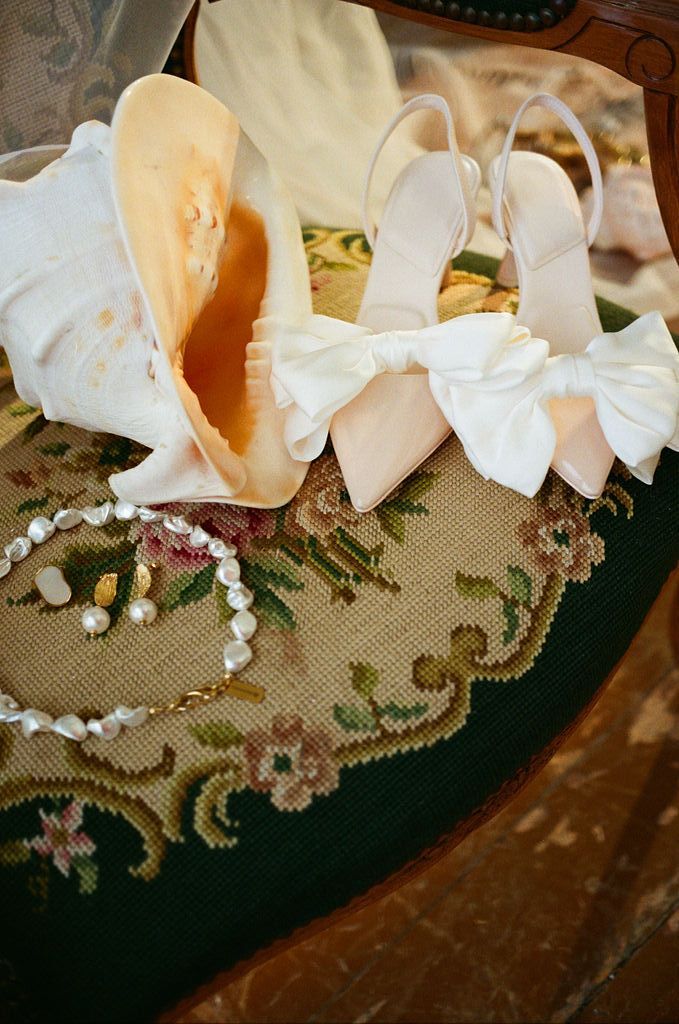 Elegant bridal heels with white satin bows beside a pearl necklace and seashell on a floral needlepoint cushion.