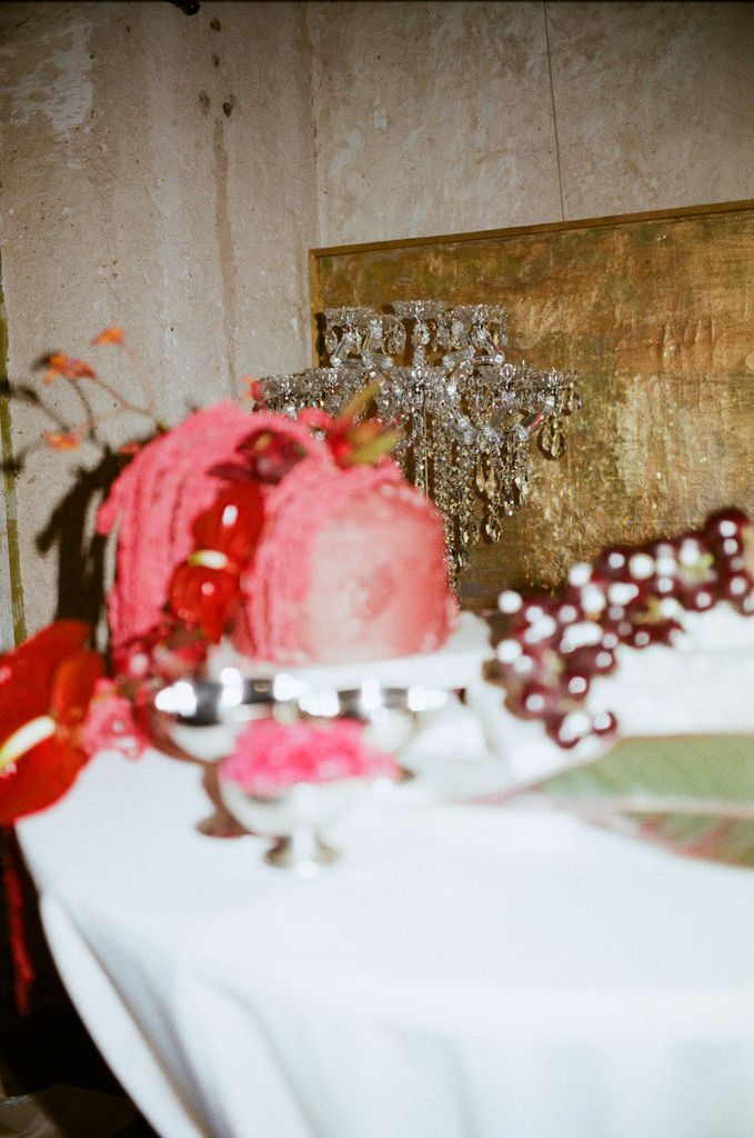 Elegant wedding reception table with pink cake, crystal candelabra, red floral arrangements, and grape clusters on white linen.