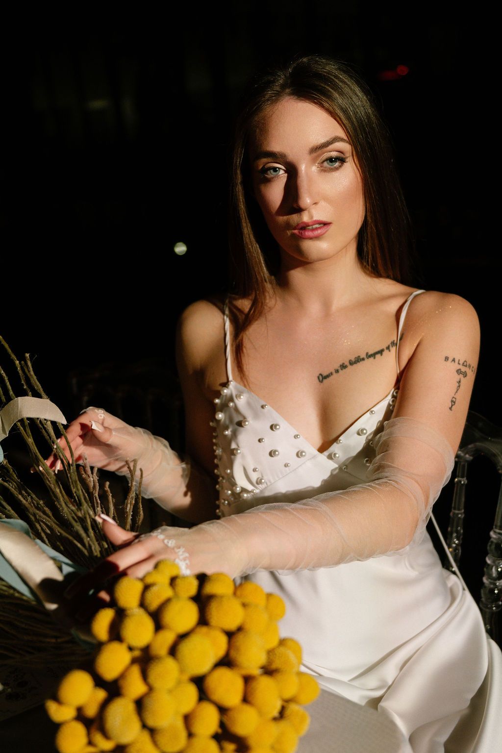 Young woman in white satin slip dress with pearl details and tattoos poses with yellow flowers in dark setting.