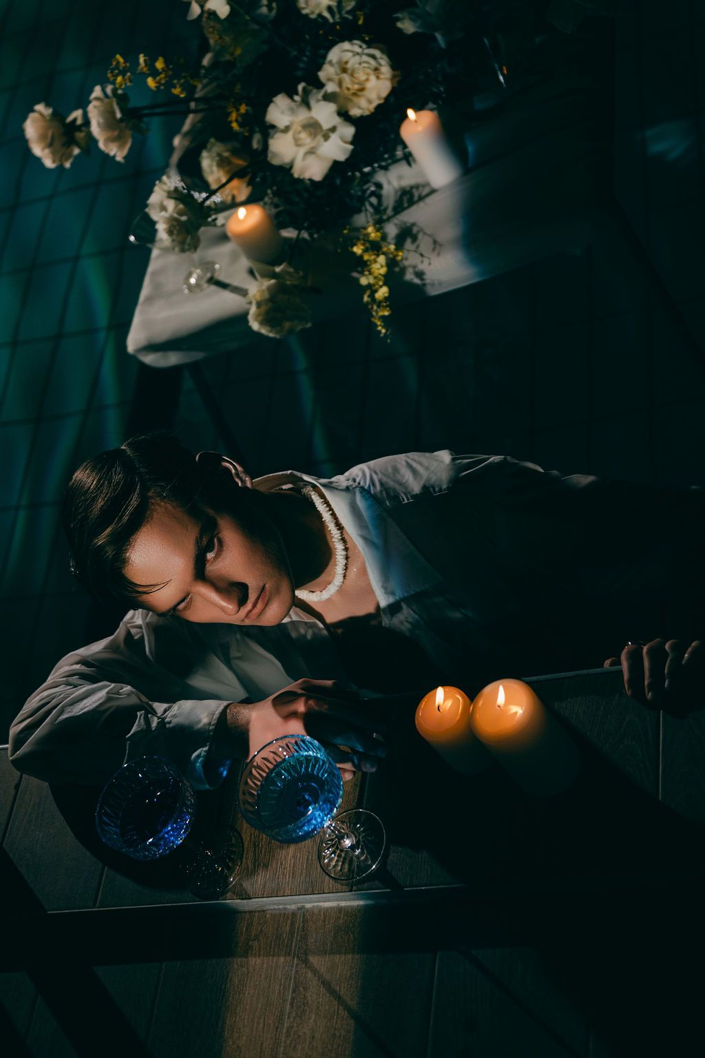 A person rests their head on a dark table surrounded by glowing candles and white flowers in dim, moody lighting.