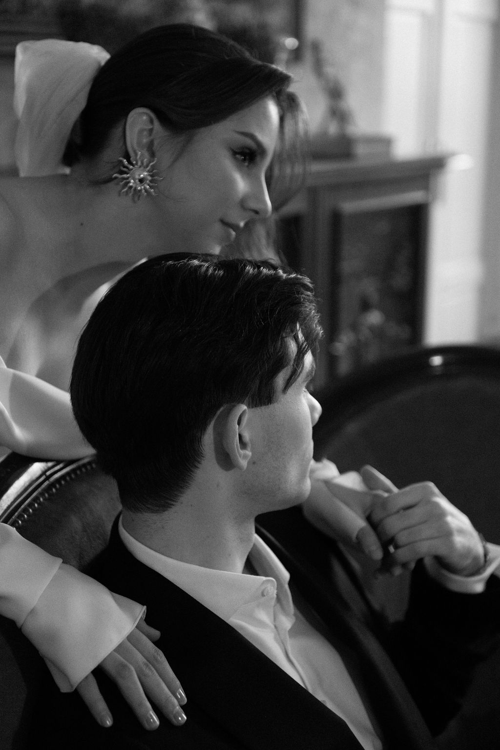 Black and white photo of an elegant couple, woman in strapless gown with star earring embracing seated man in tuxedo.