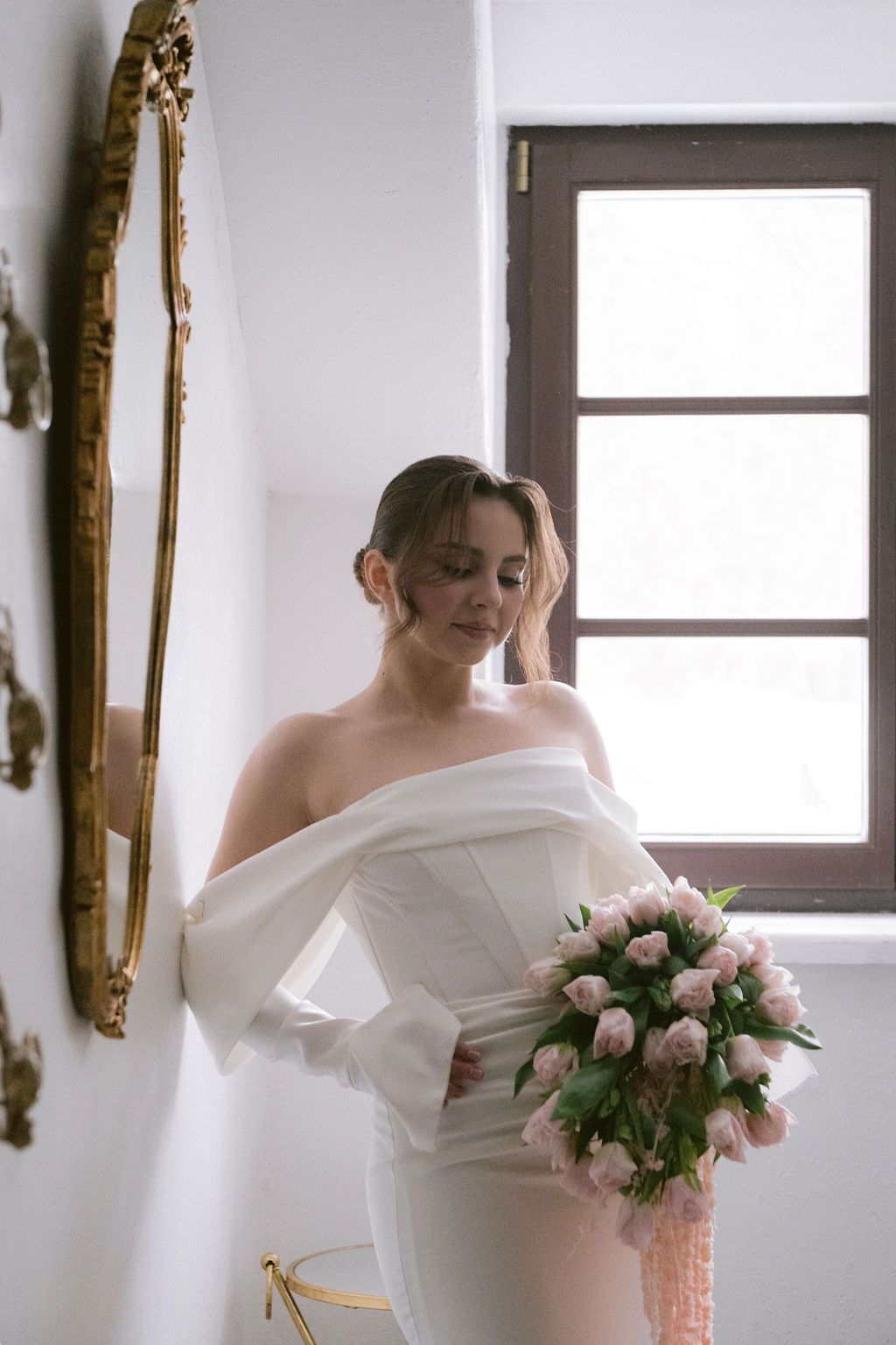 Bride in off-shoulder white gown holding pink tulip bouquet near ornate mirror and window.