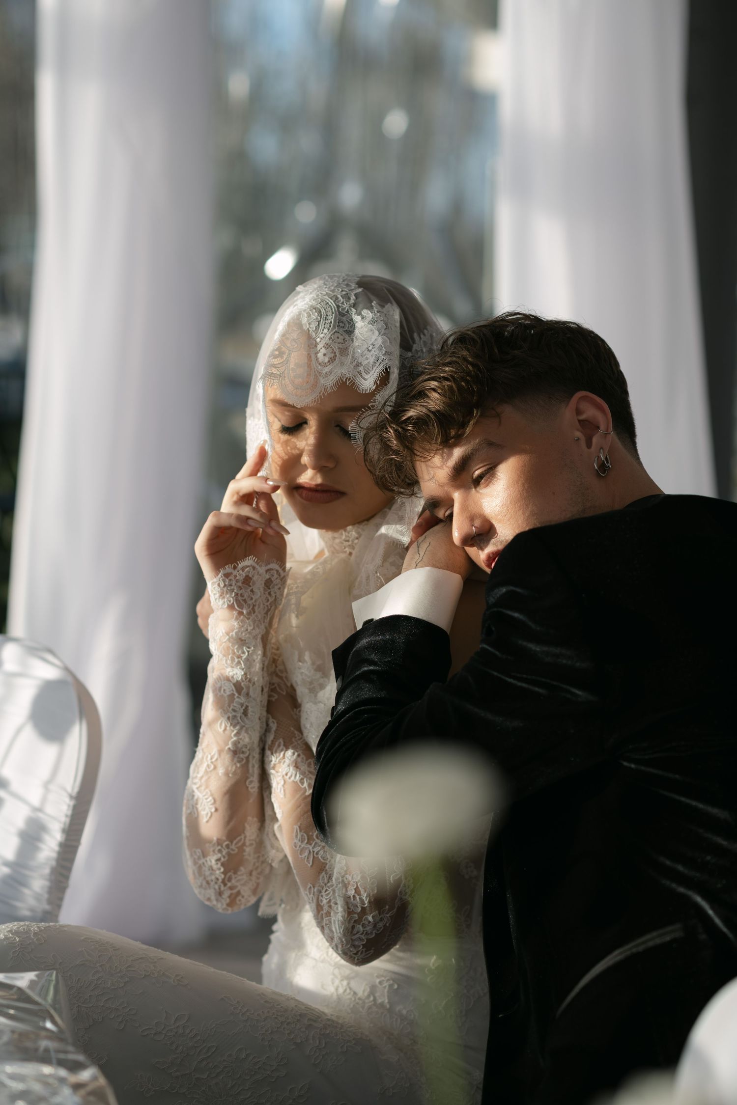 Elegant bride in lace dress and veil leans close to groom in black suit during intimate wedding portrait.