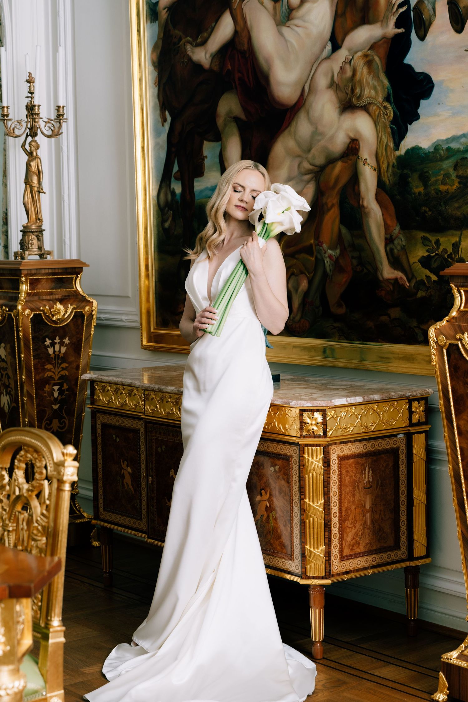Blonde bride in an elegant white satin wedding gown holding white calla lilies in an ornate room with gilded furniture and a large painting.