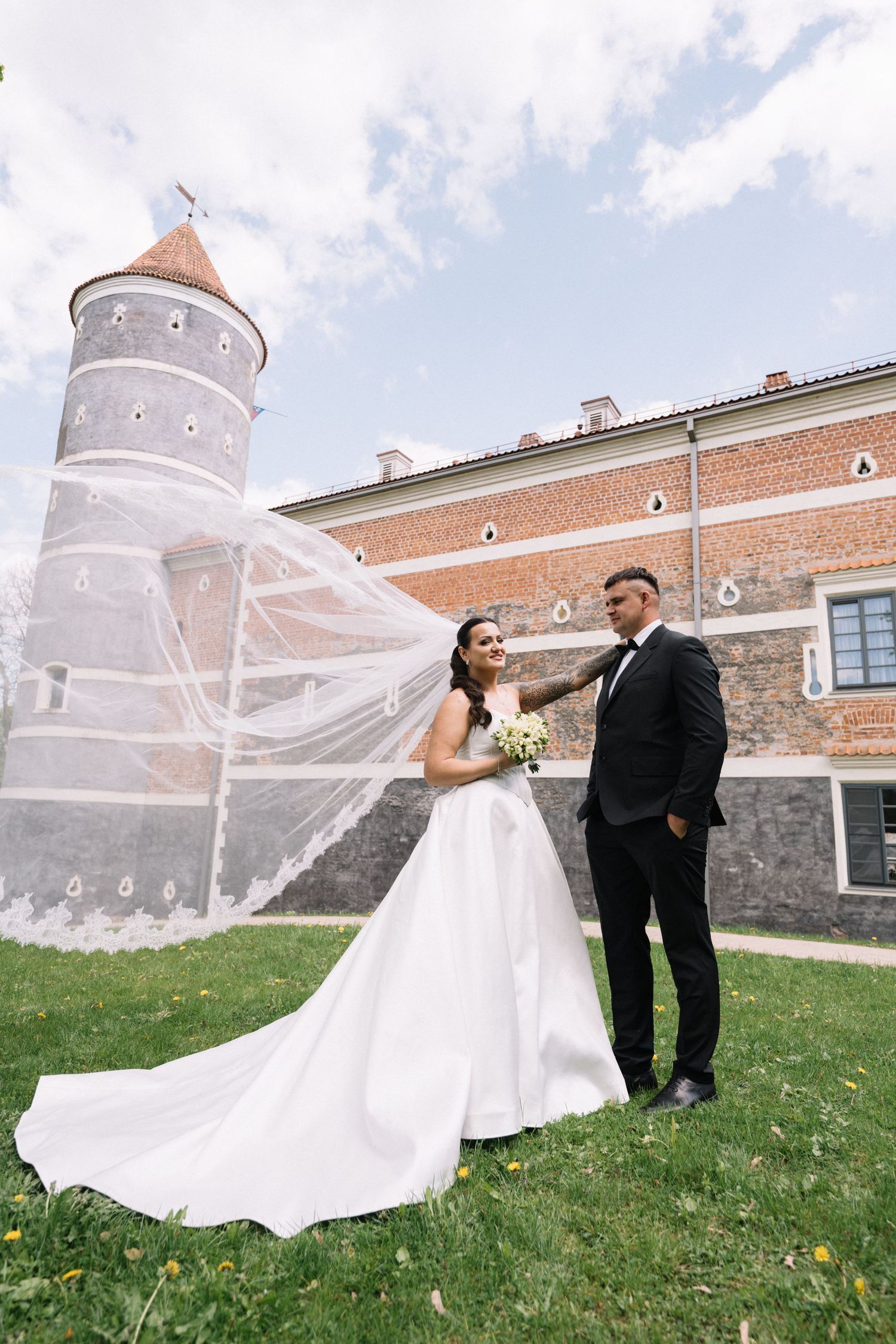Bride in a flowing white gown and veil holds a bouquet beside her groom in a black suit on a lawn before a historic brick castle tower.