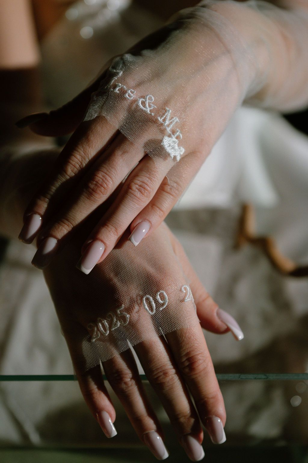 Bride's hands with long nude nails wearing sheer tulle gloves embroidered with initials and the wedding date 2025.