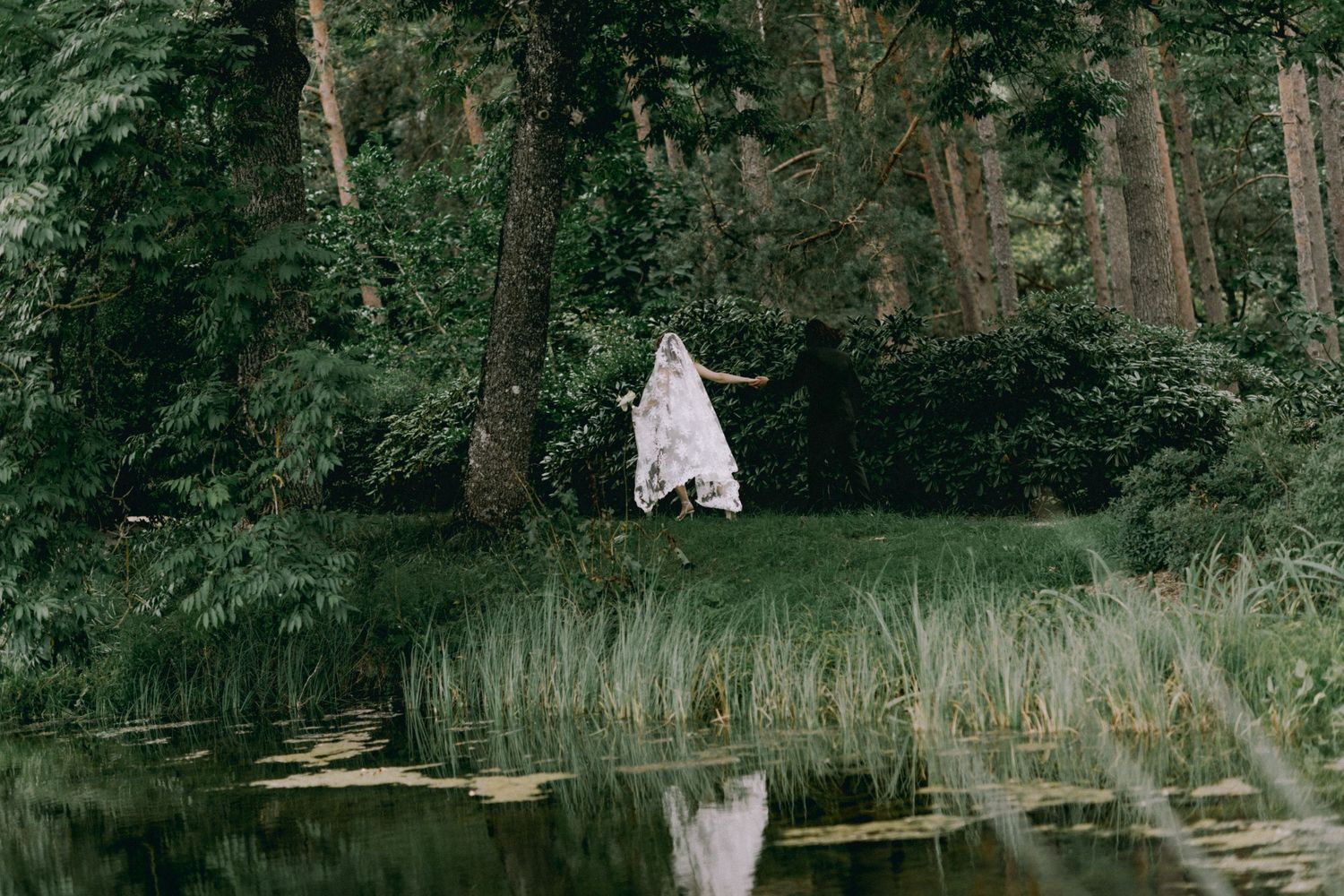 Person in a flowing white dress stands on a grassy bank beside a still pond, surrounded by tall trees in a lush green forest.