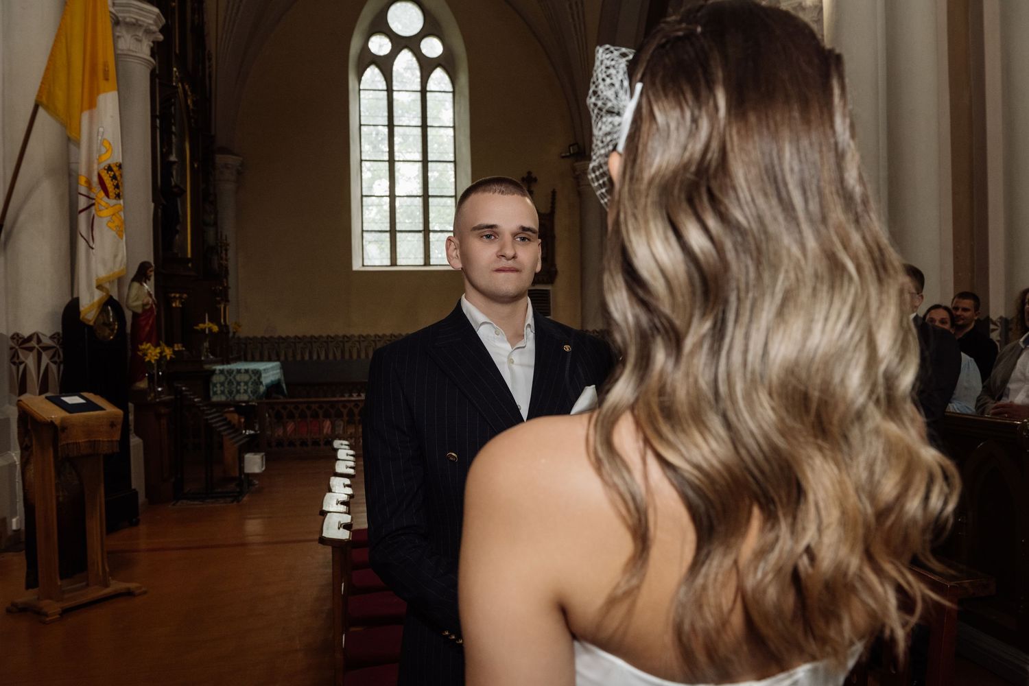 Bride with long wavy hair and a floral hair accessory faces a groom in a dark suit during a wedding ceremony inside a church.