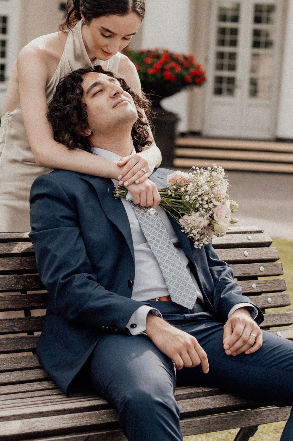 Bride stands behind groom on bench, holding bouquet, as he looks up at her lovingly in navy suit.