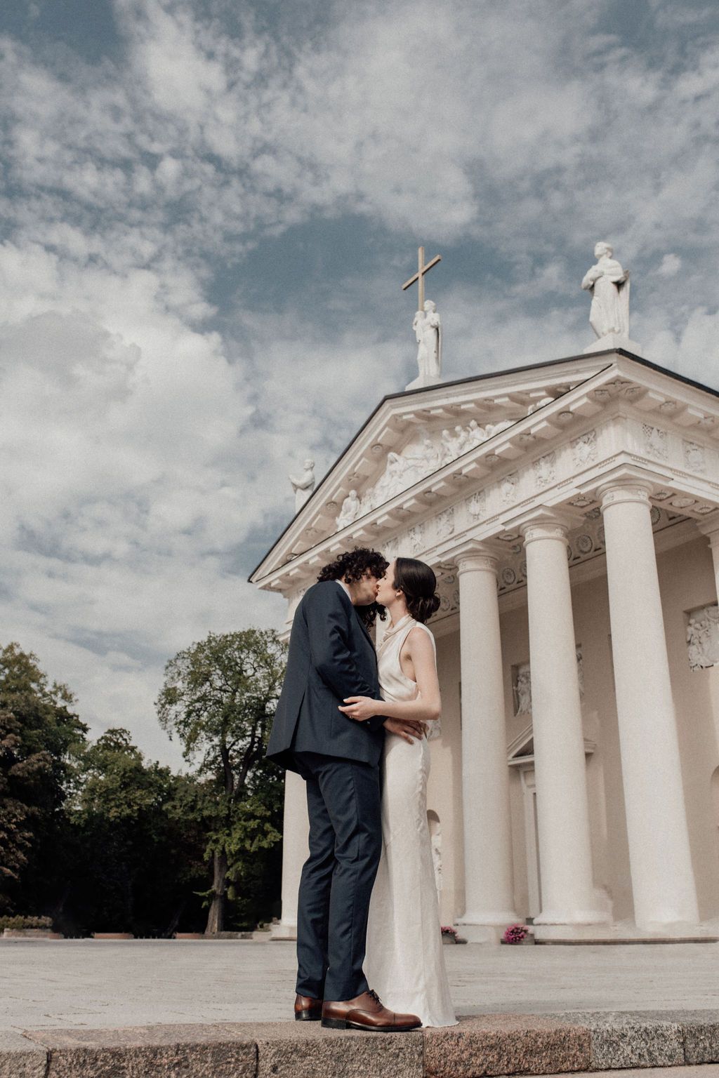 Bride and groom kissing in front of a white neoclassical church with columns and a cross under a cloudy sky.