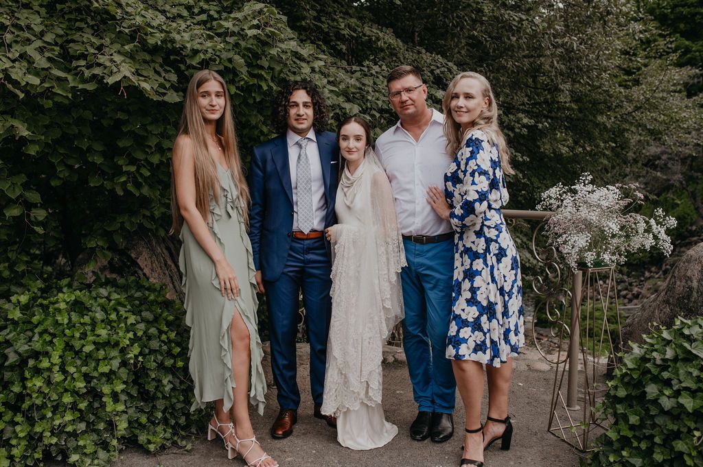 Five people posing at a wedding outdoors, bride in white dress flanked by family members smiling.