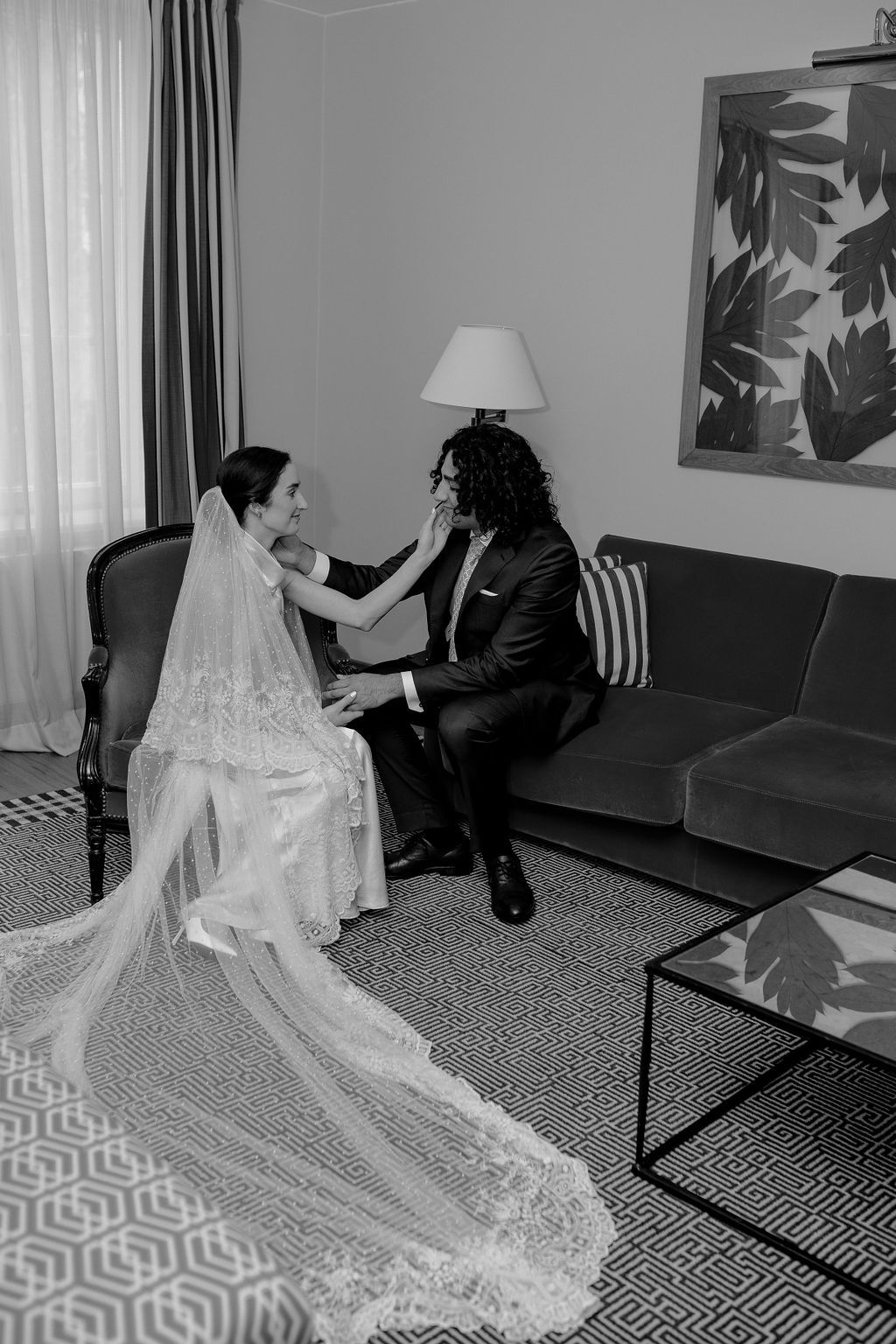 Black and white photo of a bride and groom sharing a tender moment seated in an elegant hotel room.