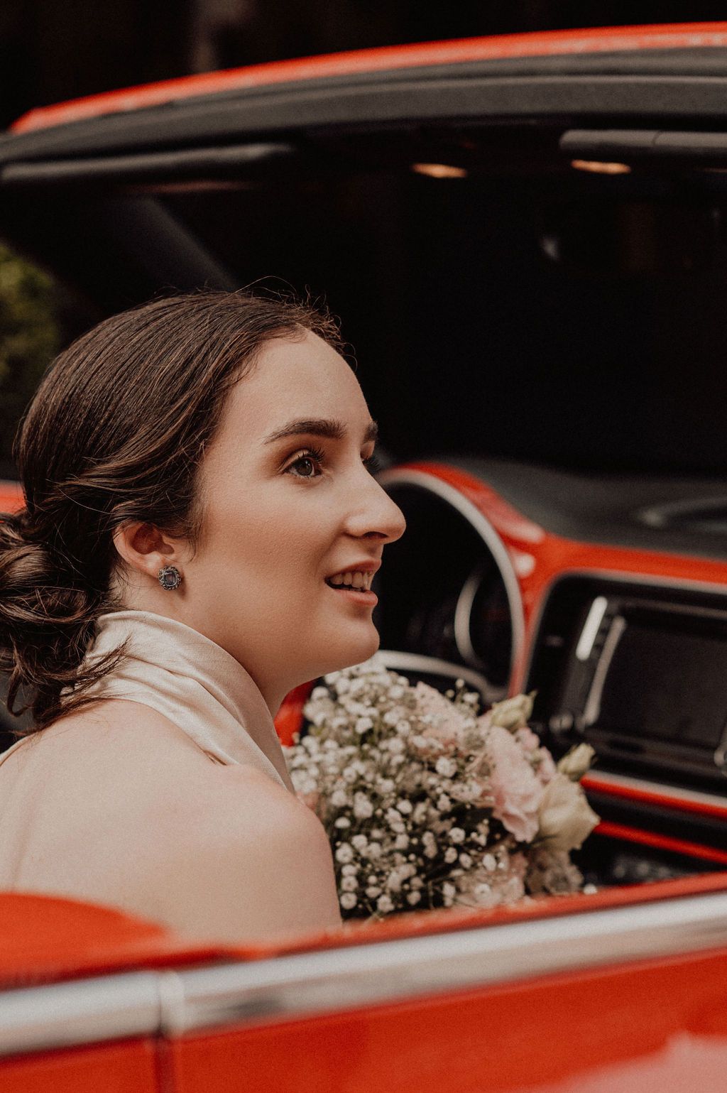 Smiling bride holding a bouquet of white and pink flowers seated in a vintage red convertible car.