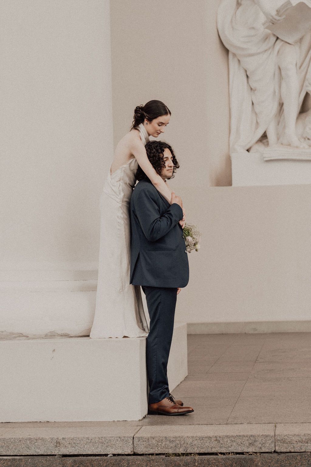 Bride in white gown embraces groom in navy suit holding bouquet, posing by classical stone columns.