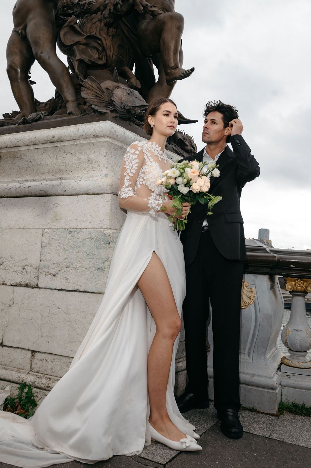 Bride in white lace gown with slit holds bouquet beside groom in black suit near ornate stone sculpture.