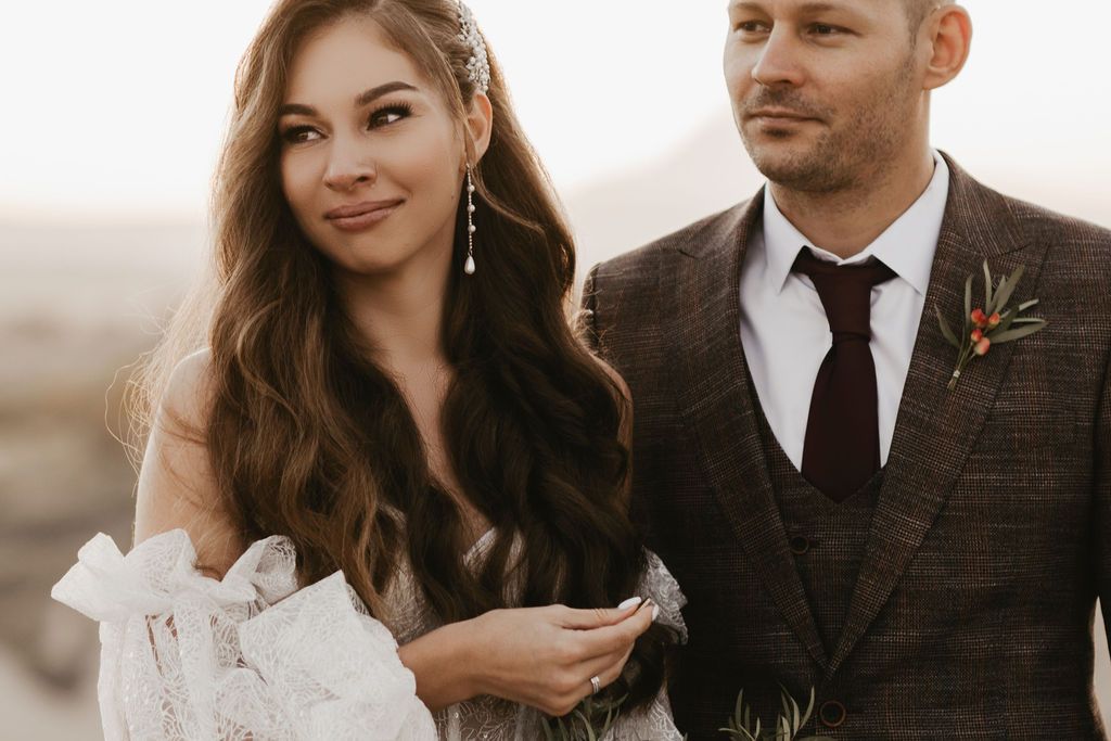 Bride and groom in elegant wedding attire pose together outdoors at golden hour, she holds white bouquet.