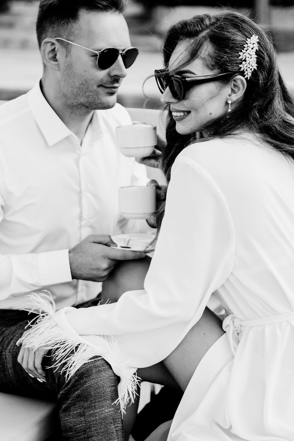 Black and white photo of a stylish couple in sunglasses sharing a romantic moment with coffee cups outdoors.