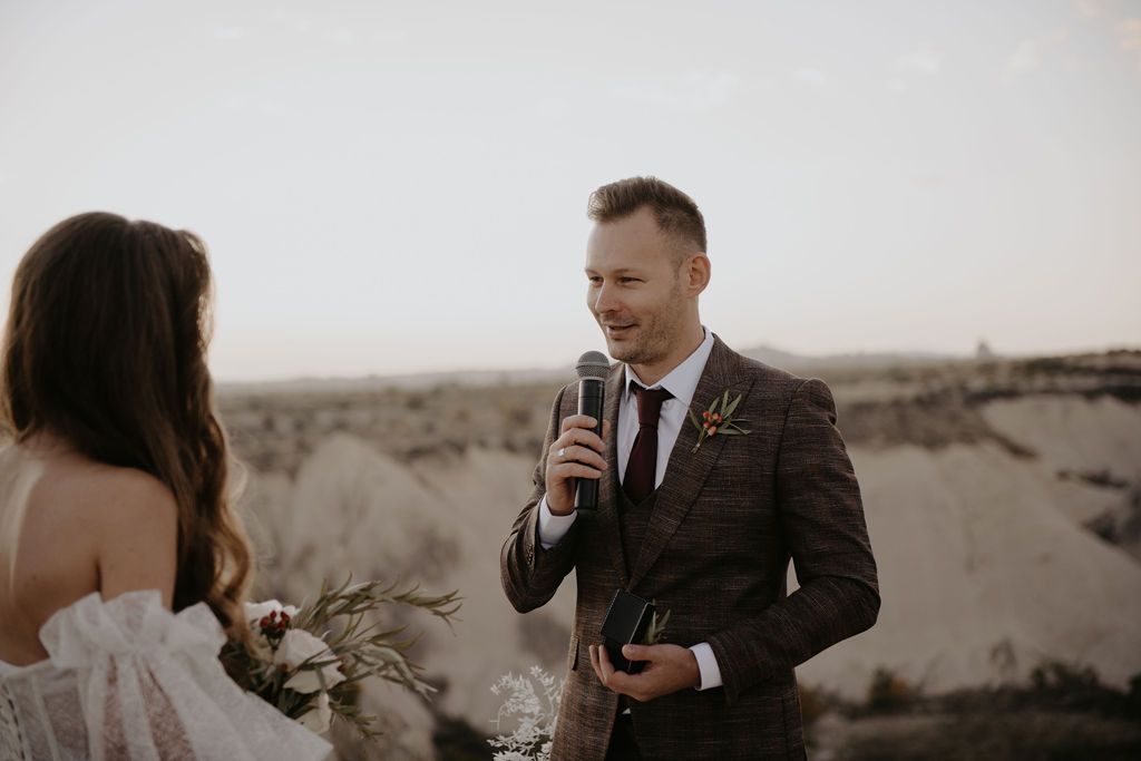 Groom in a brown suit reads vows into a microphone at an outdoor desert wedding ceremony.
