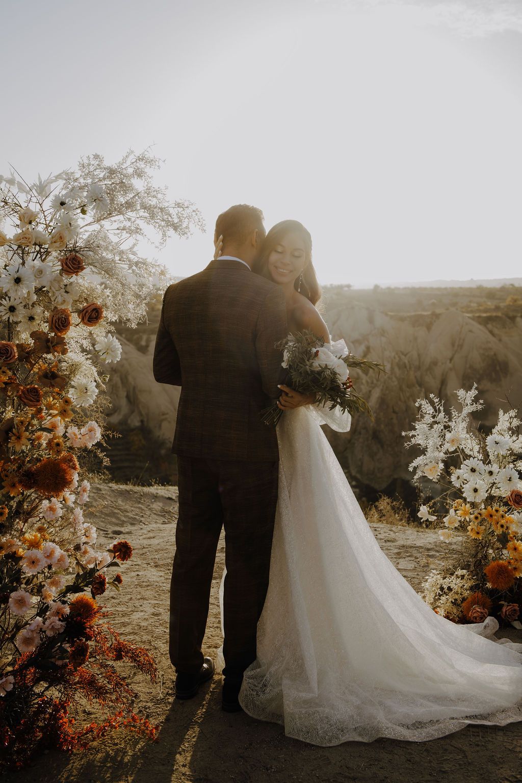 Bride and groom embrace at outdoor wedding ceremony with floral arch of white and rust blooms against a scenic desert landscape.