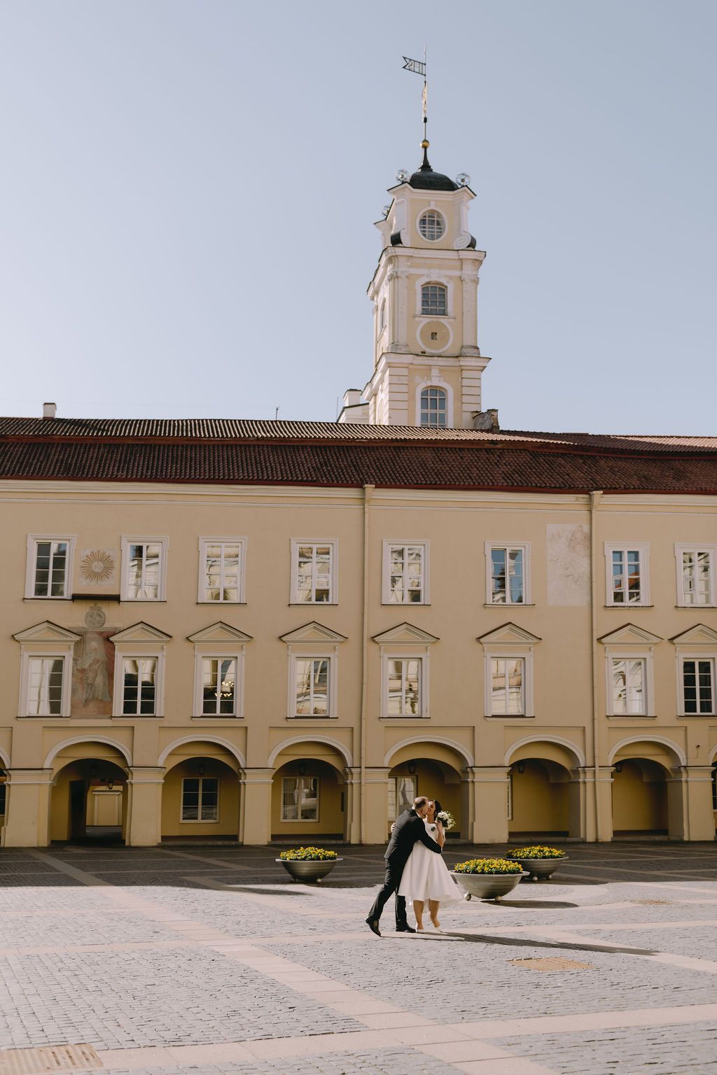 Wedding couple embracing in a sunlit European courtyard with a historic yellow building and clock tower backdrop.