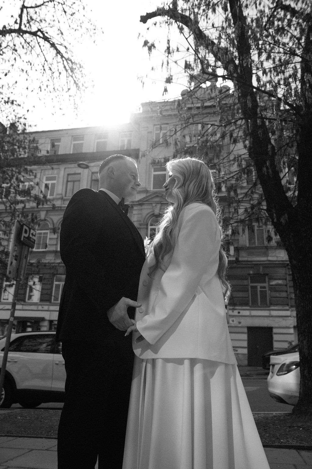 Black and white wedding photo of bride and groom holding hands outdoors, sun flare behind historic building.
