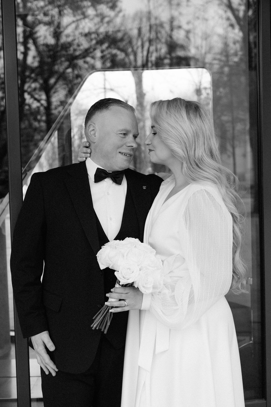 Black and white wedding photo of smiling groom in tuxedo and blonde bride in white dress holding bouquet.