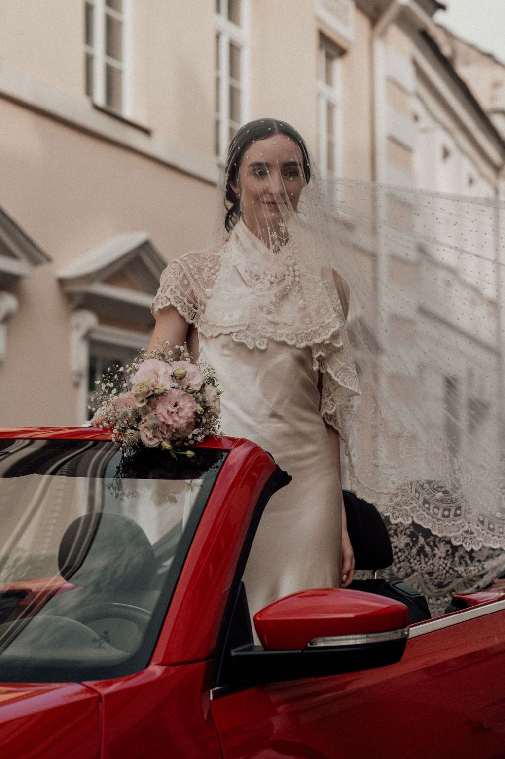 Bride in lace wedding dress and veil holds floral bouquet while standing in red convertible car on cobblestone street.